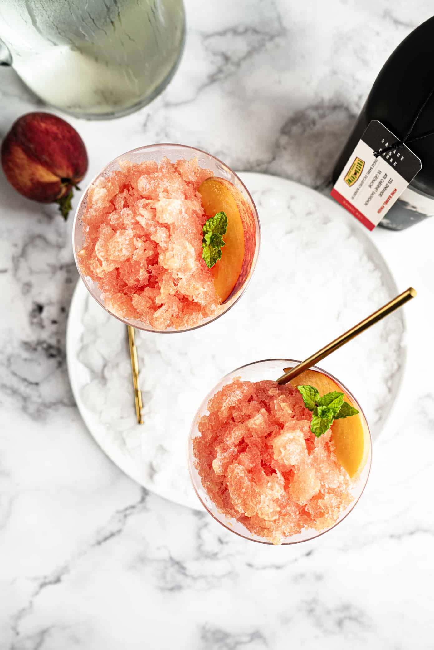 Overhead view of two glasses filled with peach slush, garnished with fresh mint and peach slices, served with gold spoons on a marble background.