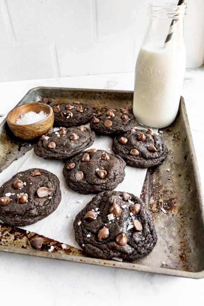 baked double chocolate chip cookies on sheet pan with milk in glass sprinkled with salt
