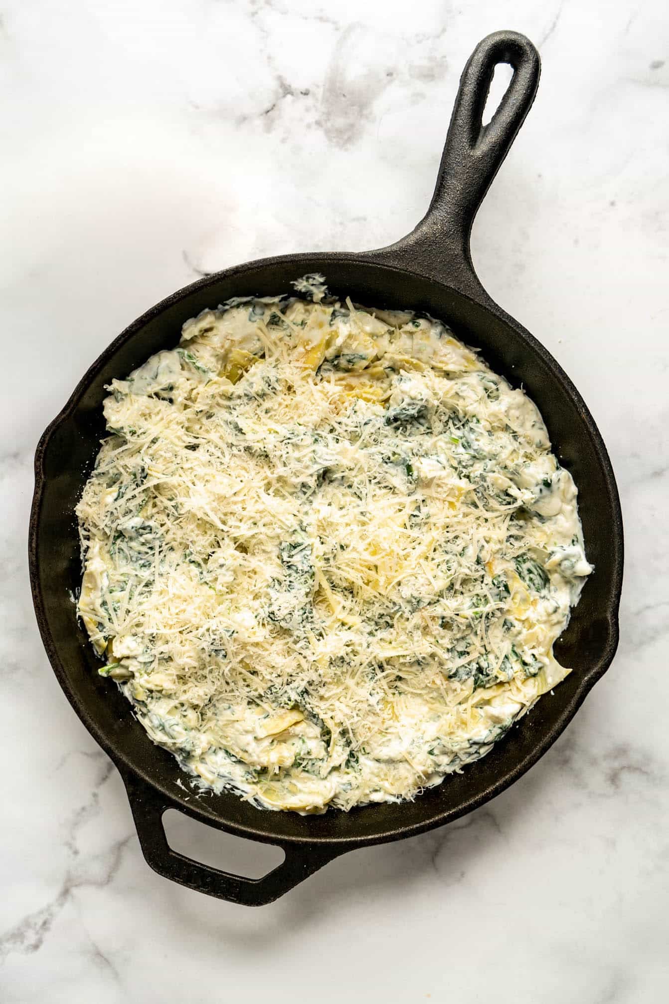 Creamy spinach and artichoke dip baked in a cast iron skillet, topped with freshly grated cheese and photographed in natural light on a marble surface.