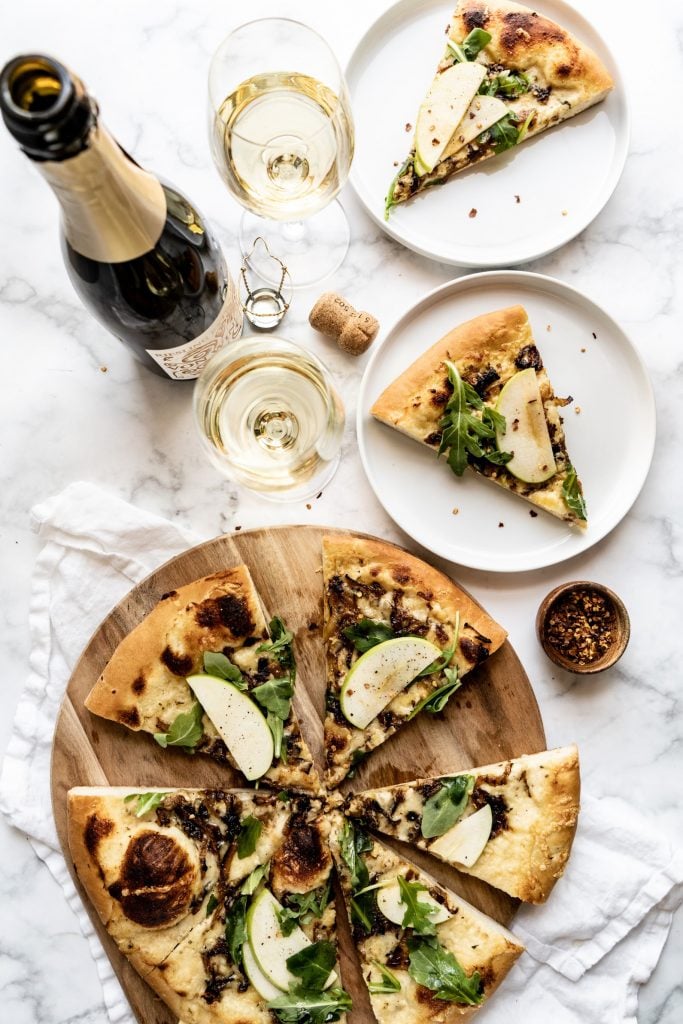 overhead shot of Gruyère and Caramelized Onion pizza on cutting board with two pieces of pizza on plates, two glasses of wine and wine bottle on table