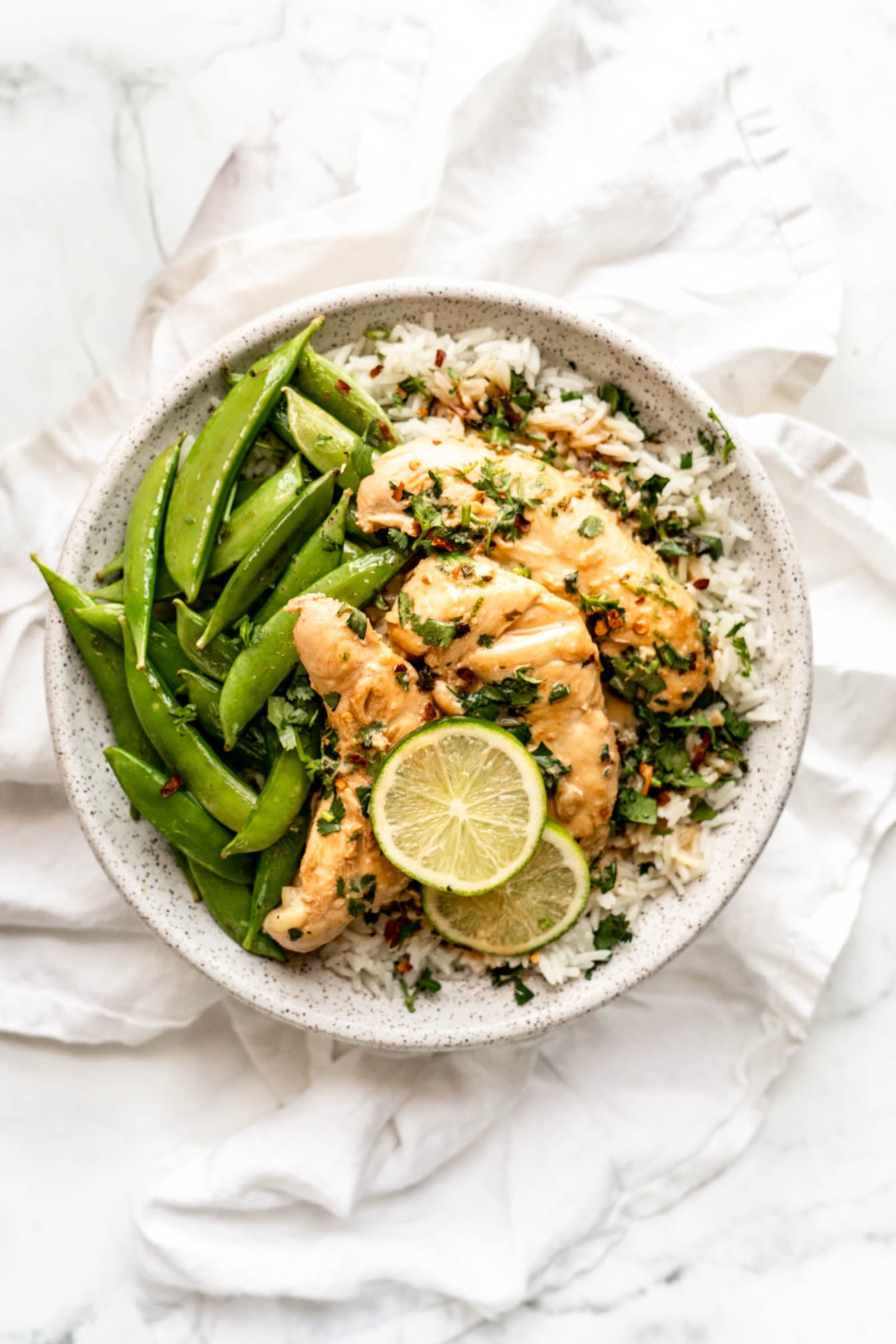 One pan coconut lime chicken served over fluffy jasmine rice with sugar snap peas, fresh cilantro, red pepper flakes, and lime slices in a light ceramic bowl on a white marble background.