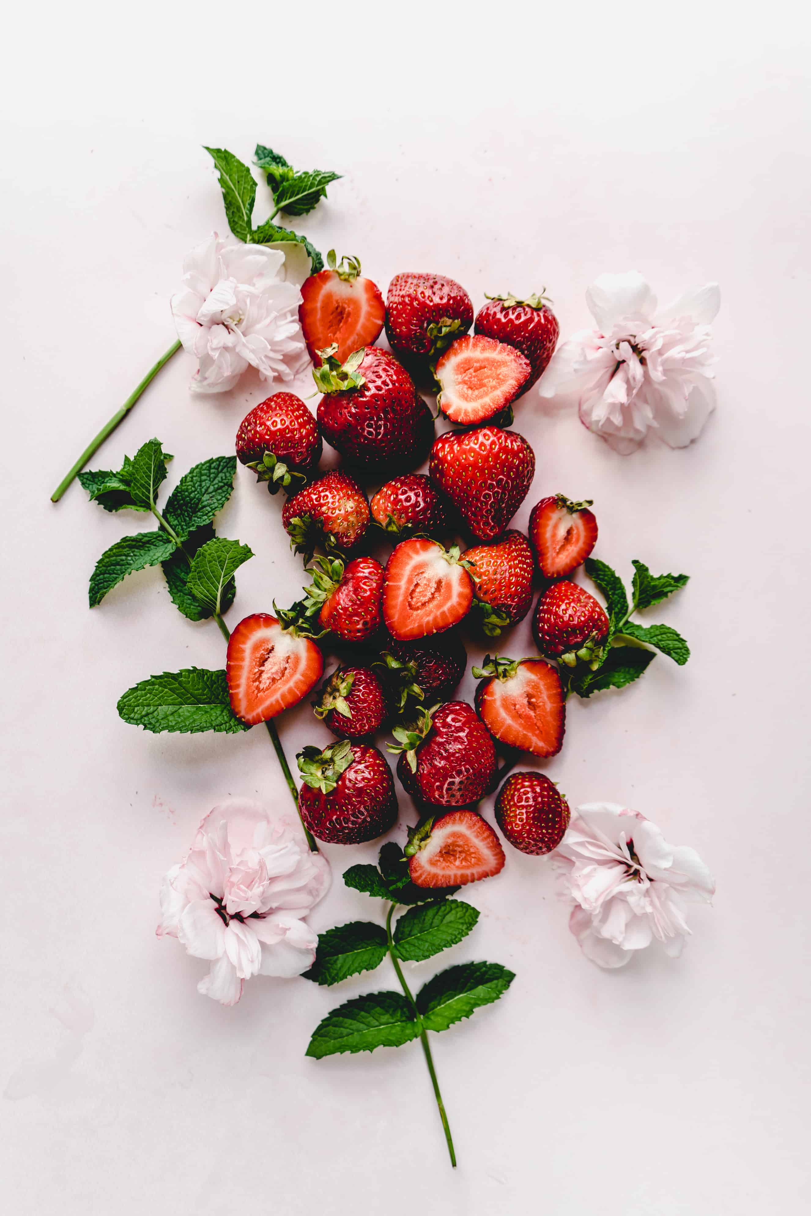 Fresh strawberries, some halved to reveal their juicy interior, are artfully arranged with mint leaves and pale pink flowers on a light pink background. This vibrant display highlights the fresh ingredients often used in strawberry bruschetta, evoking a sense of seasonal flavor and natural beauty.

