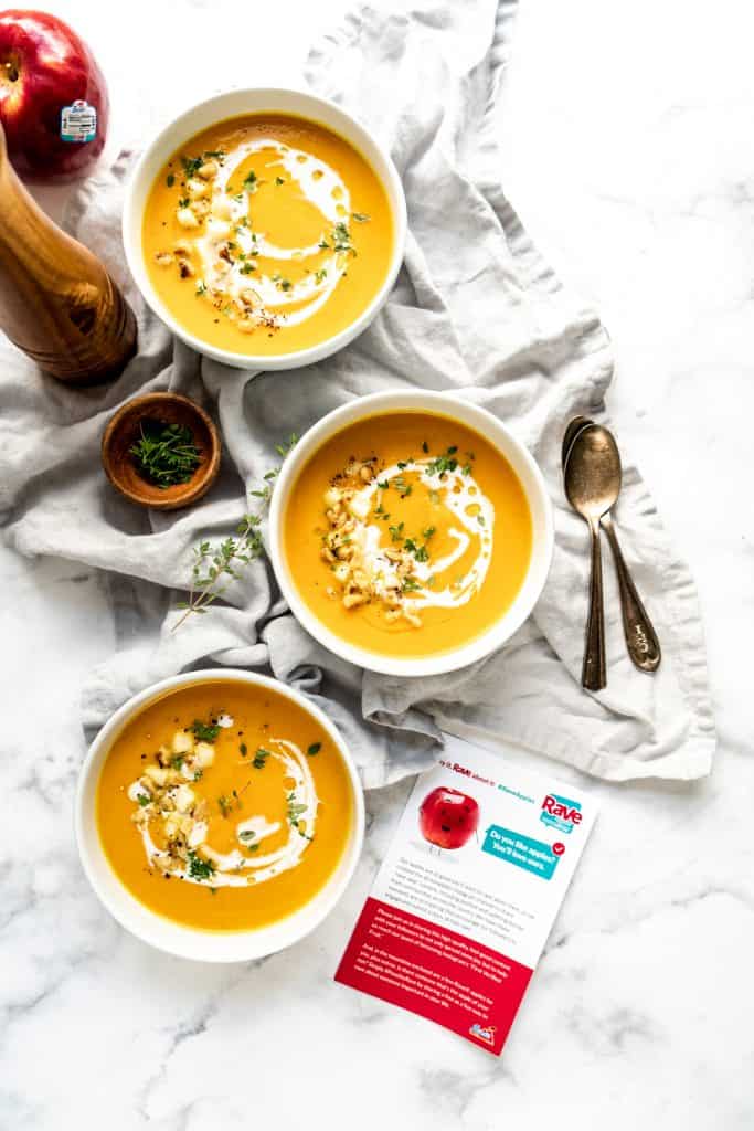 garnished carrot apple soup in three bowls with apple and pepper grinder