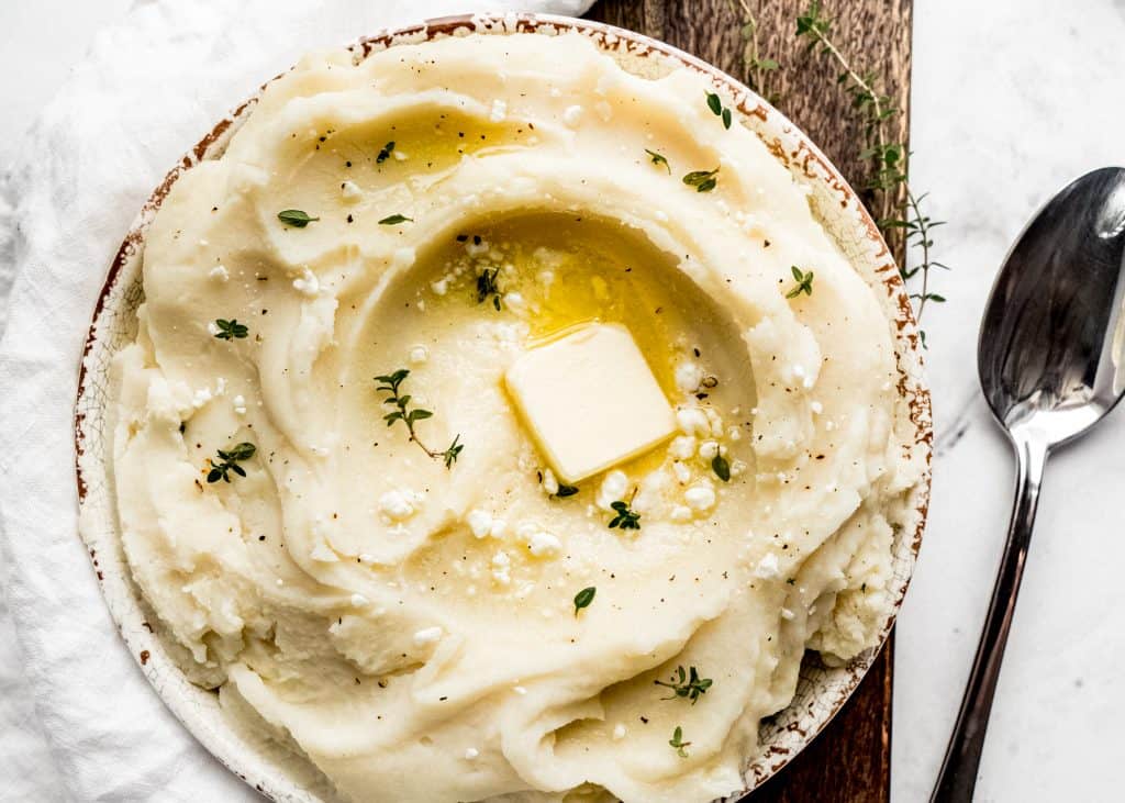 goat cheese mashed potatoes in serving bowl with butter and fresh thyme on top.  The bowl is sitting on top of a wood cutting board  with a serving spoon to the right