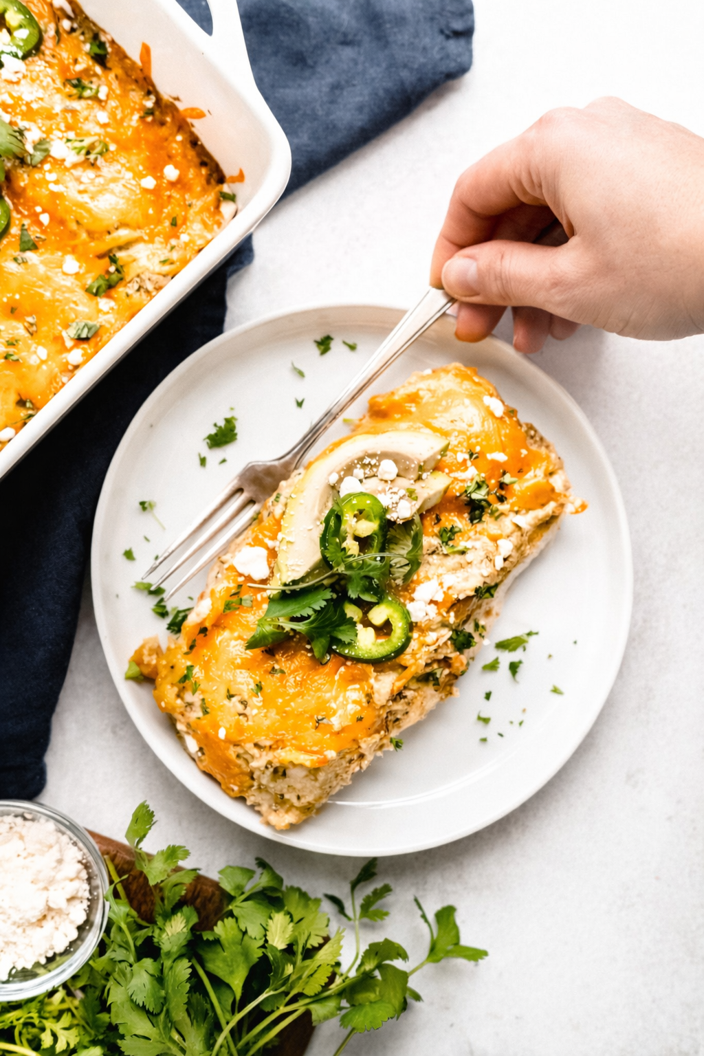 Healthy chicken enchiladas topped with melted cheddar cheese, sliced jalapeños, fresh cilantro, and avocado, served on a white plate with a hand reaching in with a fork on a light background.
