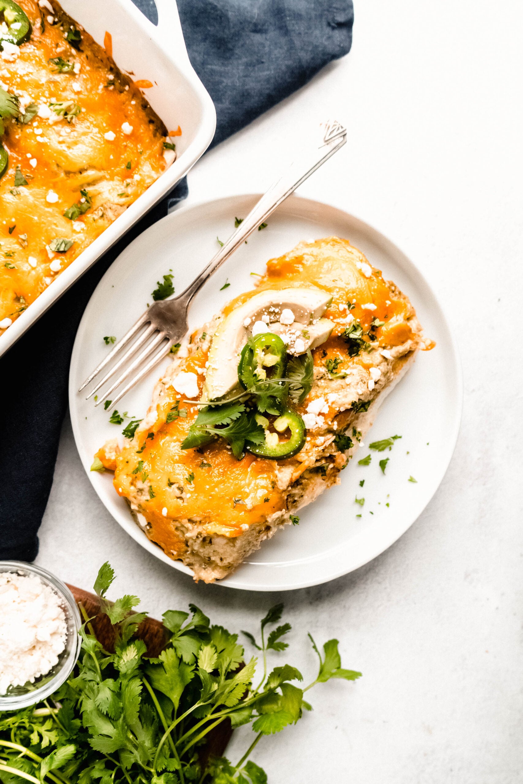 Healthy chicken enchiladas topped with melted cheddar cheese, sliced jalapeños, fresh cilantro, and avocado, served on a white plate on a light background.
