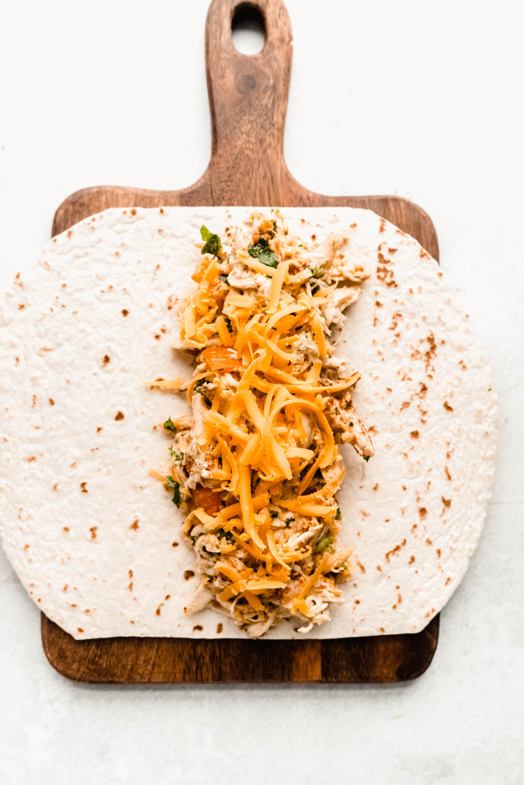 Healthy chicken enchiladas being assembled with shredded chicken, melted cheddar cheese, and cilantro filling rolled inside a flour tortilla on a wooden cutting board.