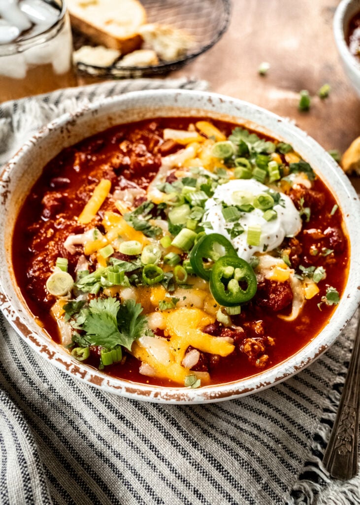 A 45 degree angle shot of a bowl of turkey chili garnished with shredded cheese, cilantro, sliced green onion and sour cream.  The bowl is sitting on a striped napkin on a wood table. 