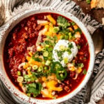 Turkey chili garnished with shredded cheese, cilantro, green onion and sour cream in a white bowl with a striped napkin underneath.