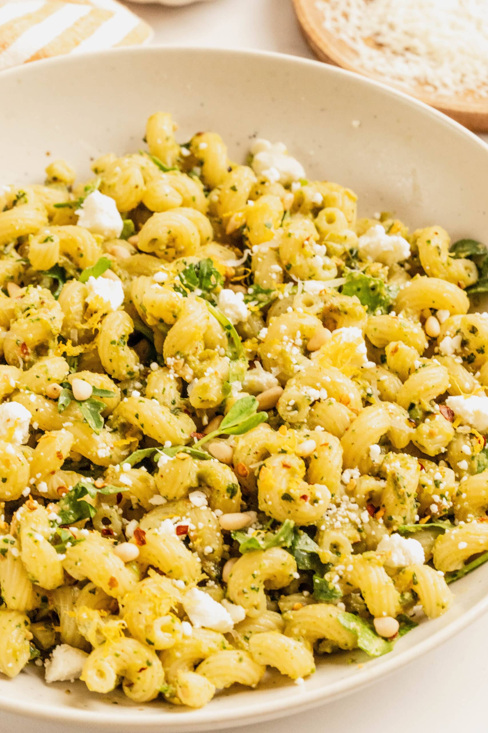 Close-up of pesto goat cheese pasta made with cavatappi, tossed in vibrant green pesto and arugula, topped with crumbled goat cheese, toasted pine nuts, red pepper flakes, and a sprinkle of lemon zest in a shallow bowl.