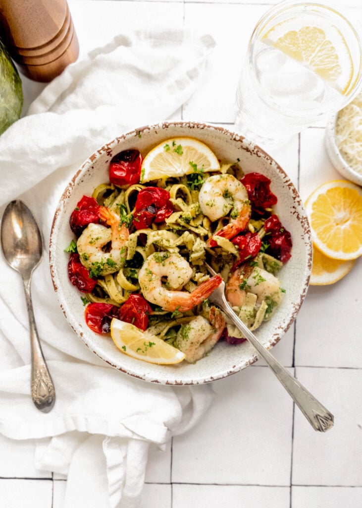Overhead view of pesto pasta with shrimp, roasted cherry tomatoes, and fettuccine in a rustic bowl, garnished with lemon wedges and fresh herbs on a white tile surface.