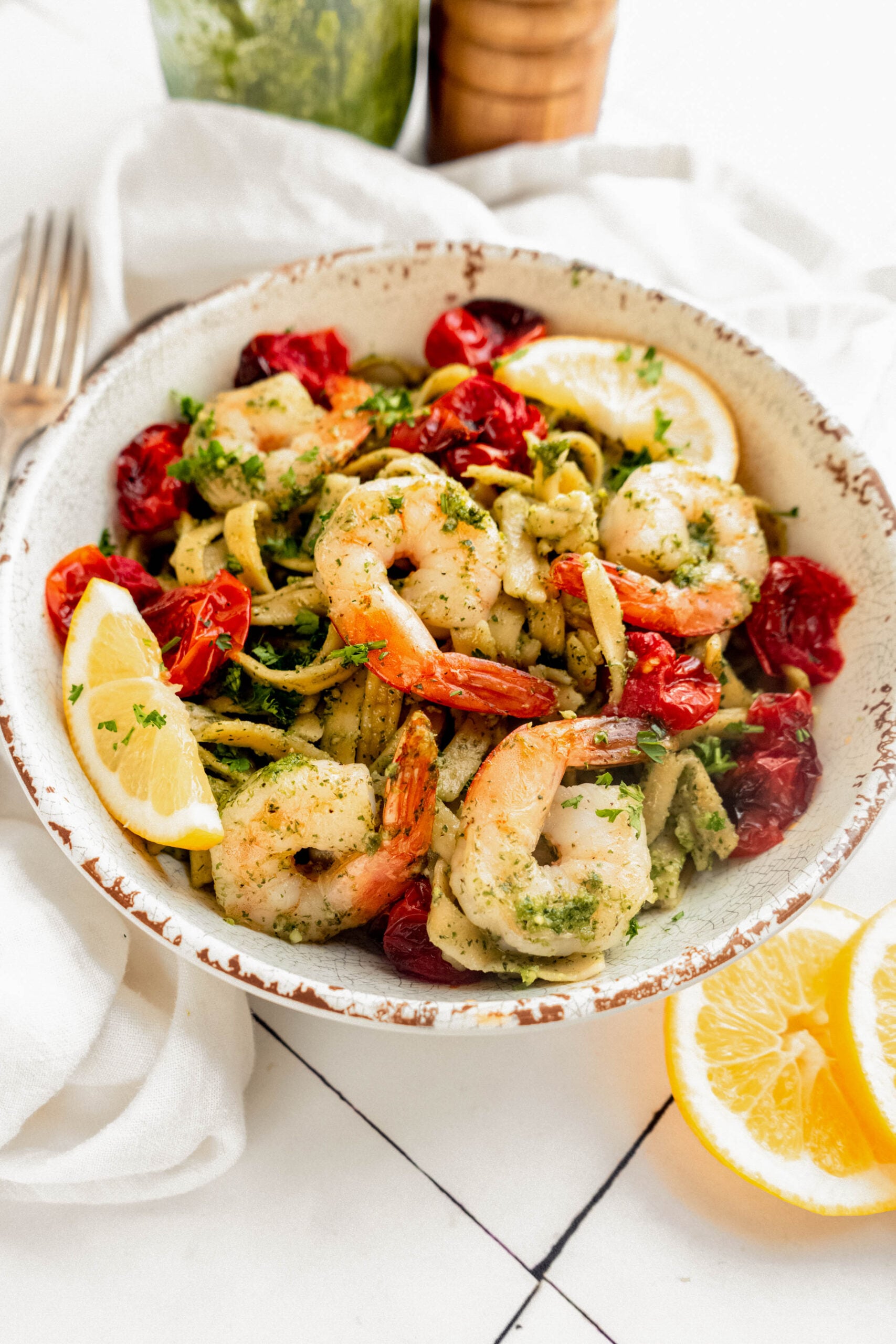 Close-up of pesto pasta with shrimp and roasted cherry tomatoes in a rustic bowl, finished with lemon wedges, fresh herbs, and parmesan on a bright white surface.