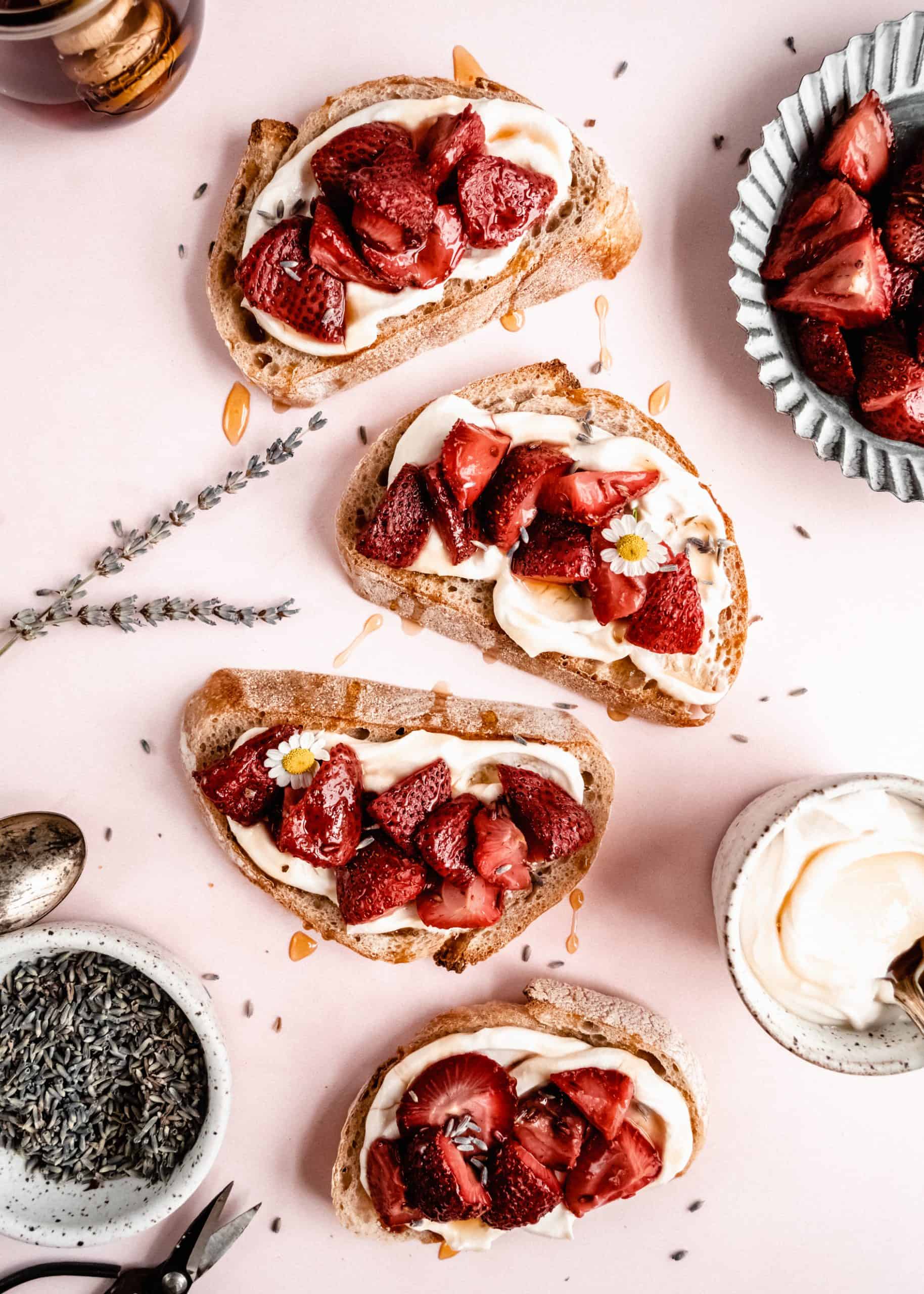 Flat lay image of strawberry bruschetta arranged on a soft pink surface. Slices of crusty bread are topped with creamy ricotta, roasted strawberries, a drizzle of honey, and garnished with chamomile flowers and lavender. Surrounding the bruschetta are ingredients and utensils, creating a fresh and elegant presentation.
