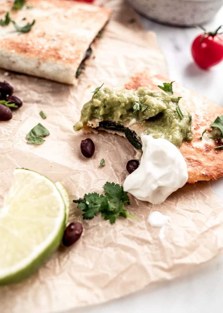 closet up shot of bite taken out of poblano and black bean quesadillas dipped in guacamole and sour cream laying on parchment paper