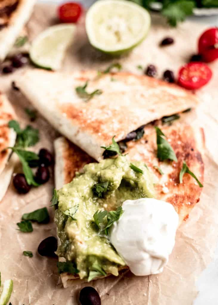 closet up shot of poblano and black bean quesadillas dipped in guacamole and sour cream laying on parchment paper