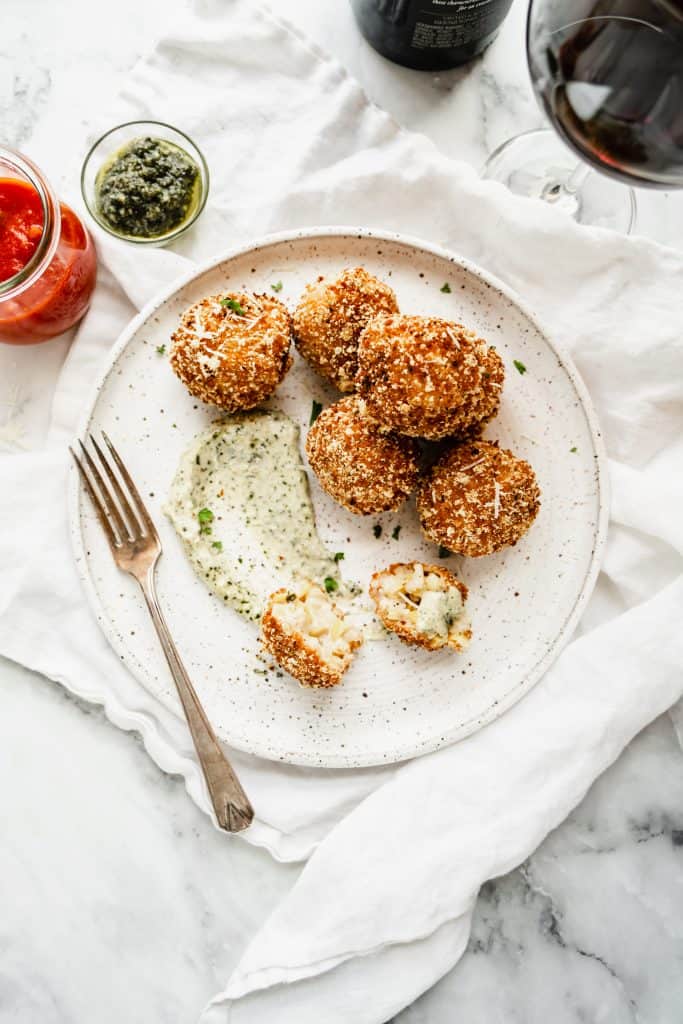 sweet corn arancini on white plate with pesto aoili spread on plate, red wine in glass and marinara and pesto in dishes on side 