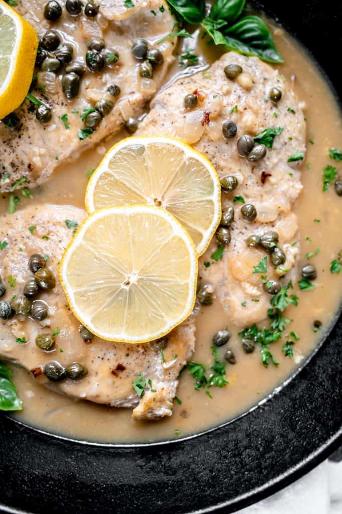 close up overhead shot of Chicken piccata garnished with lemon slices, basil and parsley in a cast iron pan.
