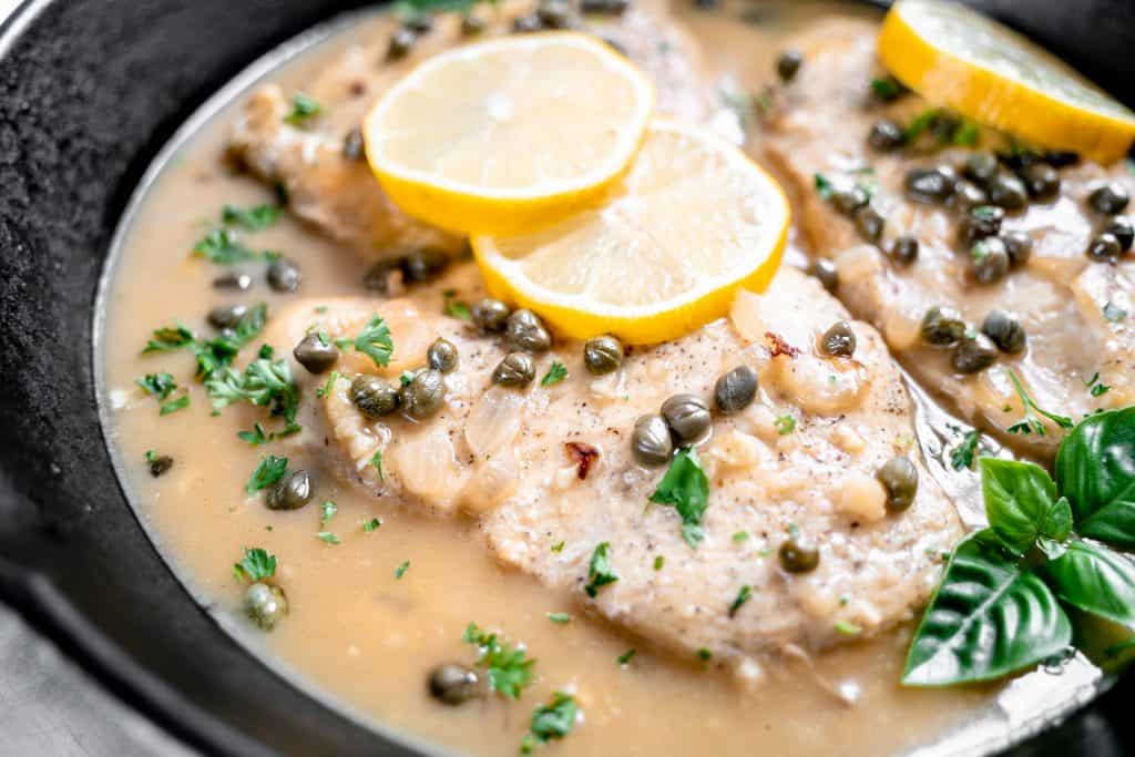 Chicken piccata garnished with lemon slices, basil and parsley in a cast iron pan.