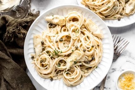 canned artichoke and lemon pasta plated on two round dishes surrounded with a dark green napkin, a glass of wine and a sliced lemon