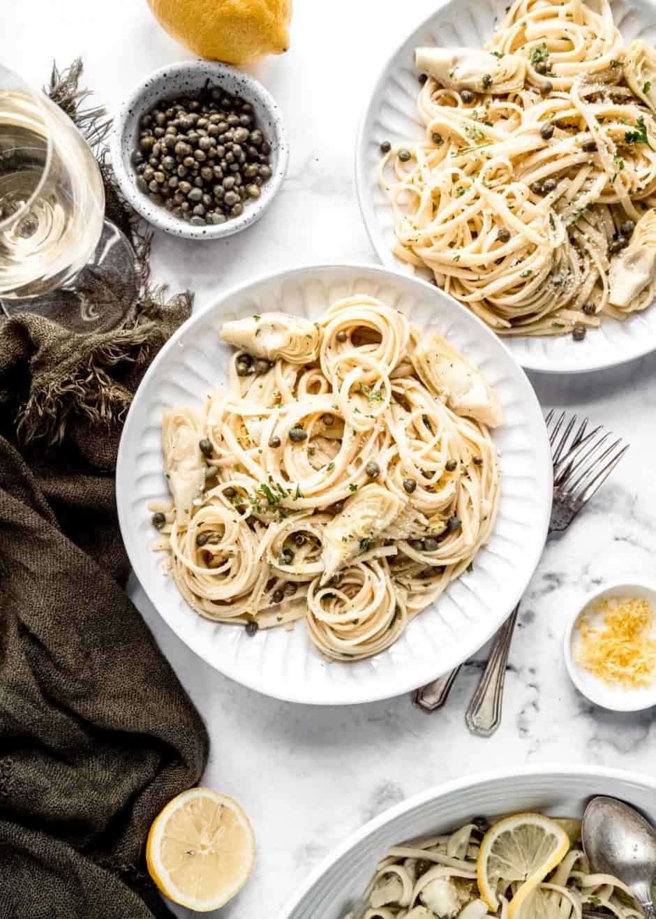 canned artichoke and lemon pasta plated on two round dishes surrounded with a dark green napkin, a glass of wine and a sliced lemon