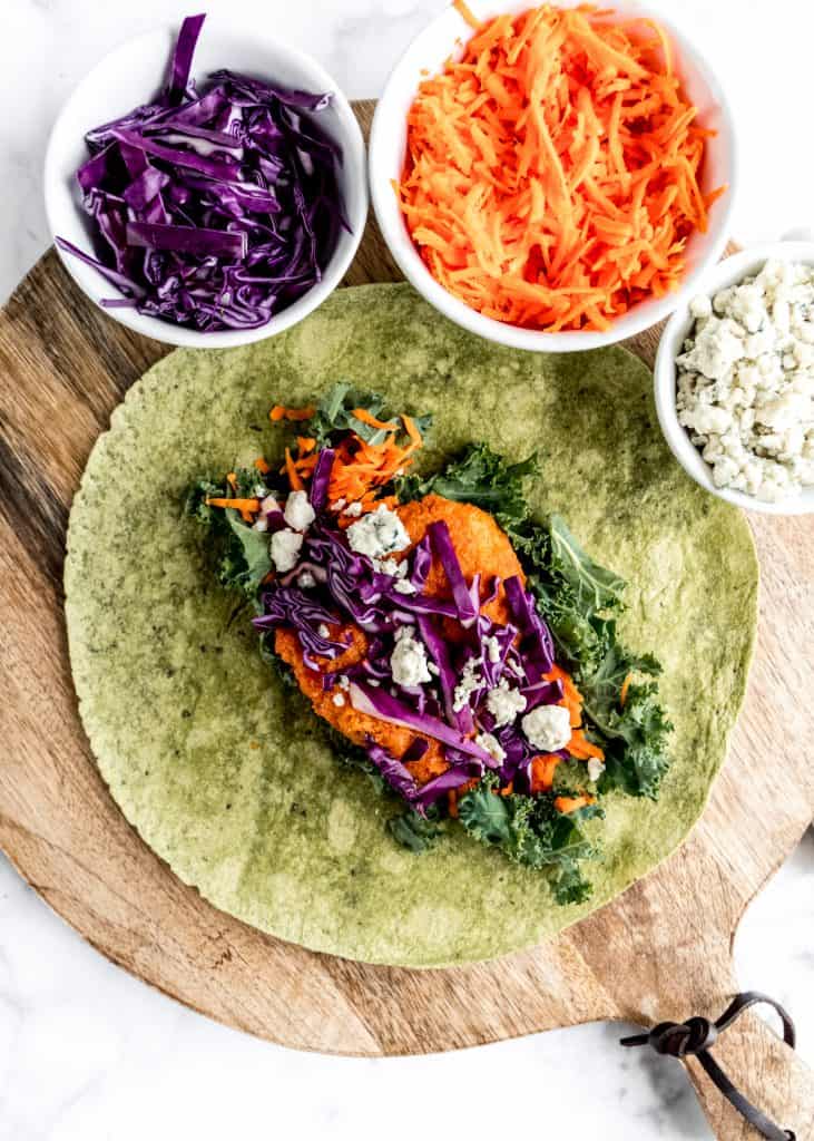 healthy buffalo chicken wrap open on cutting board with toppings in bowls around wrap