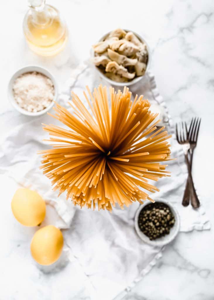 top view of linguine pasta, lemons, capers, parmesan and artichoke hearts on marble background