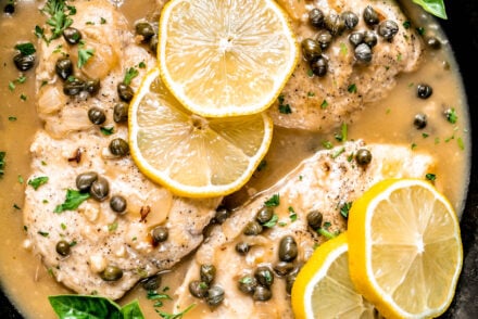 Healthy chicken piccata recipe made with pan-seared chicken breasts simmered in a lemon garlic white wine sauce with capers and shallots, garnished with fresh basil and lemon slices in a cast iron skillet.