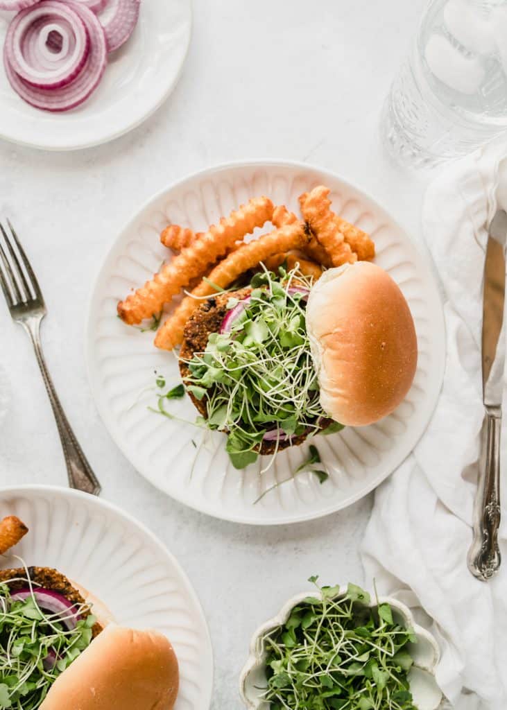 overhead shot of sweet potato black bean burger on plate topped with red onion and microgreens and served wwith fries on plate