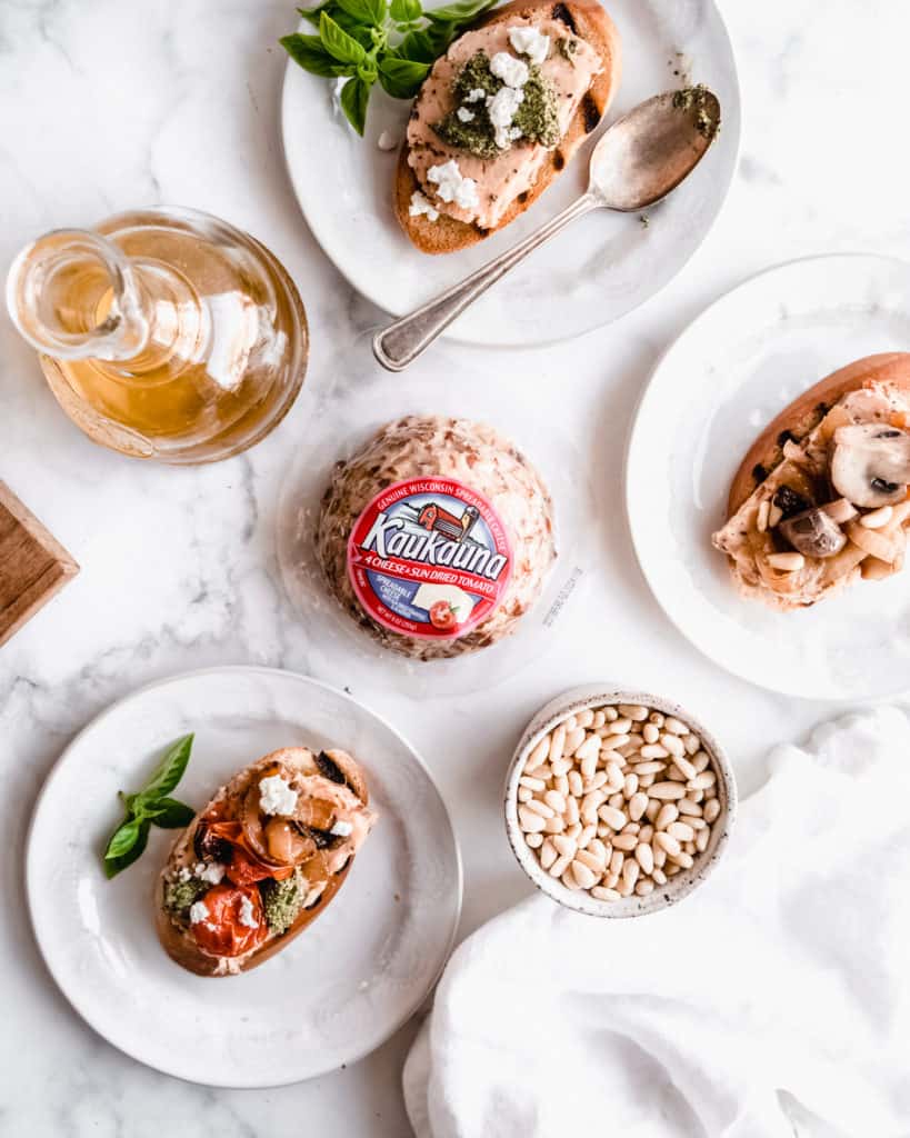 Three plates with assembled bruschetta with various toppings on plates, kaukauna cheese ball in the middle and a bottle of olive oil all on the marble background