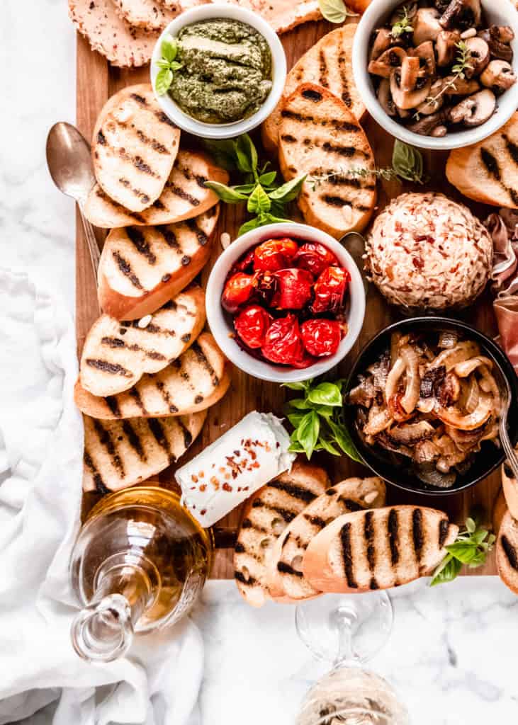 close up of pesto, sauteed mushrooms, and roasted grape tomatoes on board in bowls with grilled bread surrounding 