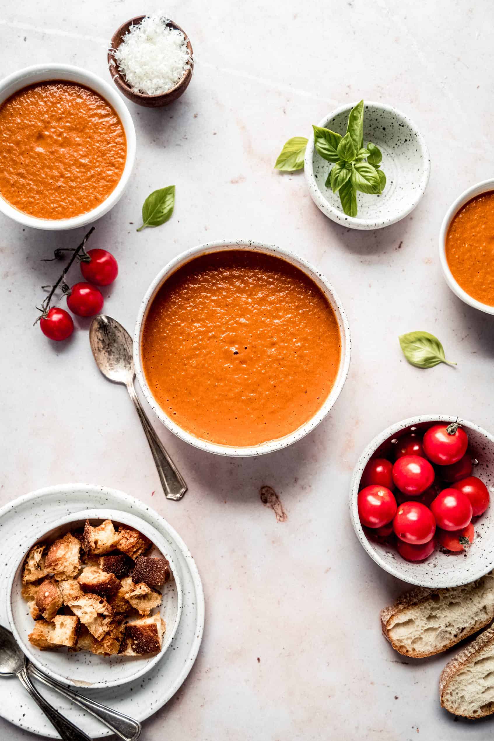 Roasted tomato soup in bowls with smaller bowls of fresh basil, parmesan cheese, cherry tomatoes and croutons