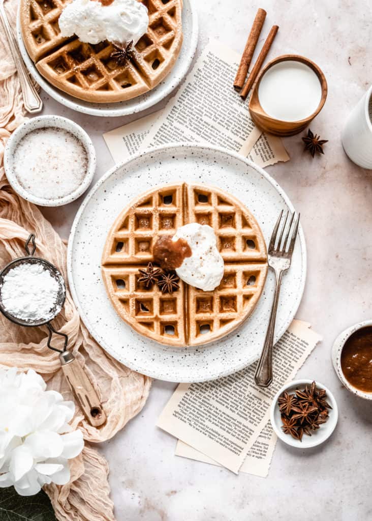 apple butter waffles garnished with cinnamon whipped cream and apple butter on plate next to cup of coffee and maple syrup in wood cup