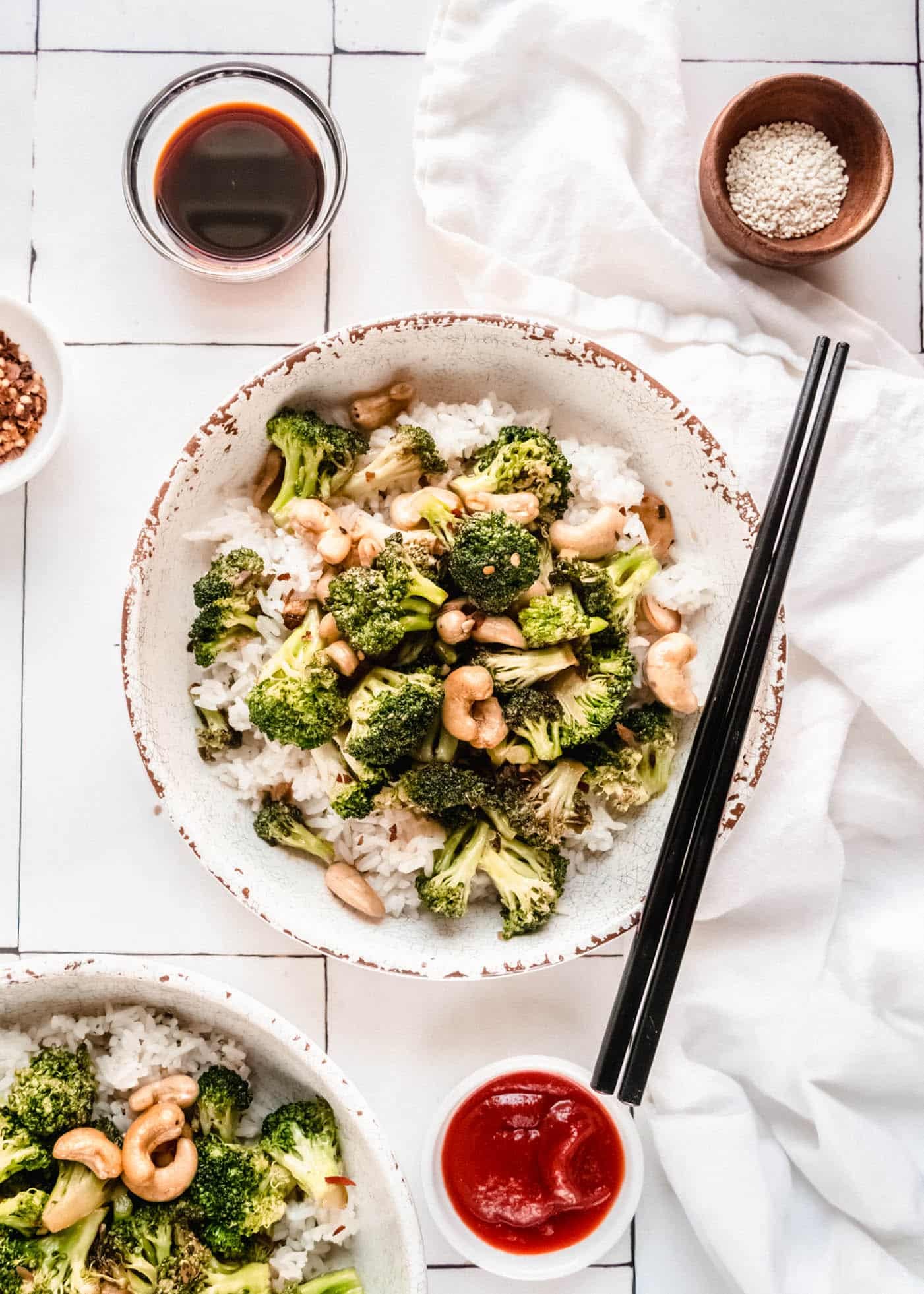 Overhead view of broccoli cashew stir fry served over white rice in rustic bowls, featuring tender broccoli florets, toasted cashews, and a savory glossy sauce with chopsticks and sauce bowls on the side.