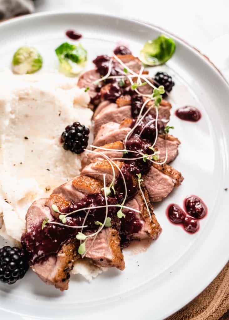 pan seared duck breast with blackberry sauce poured over top, fresh blackberries on the plate and mashed potatoes on the side.  Green brussels sprout leaves are on the plate. 