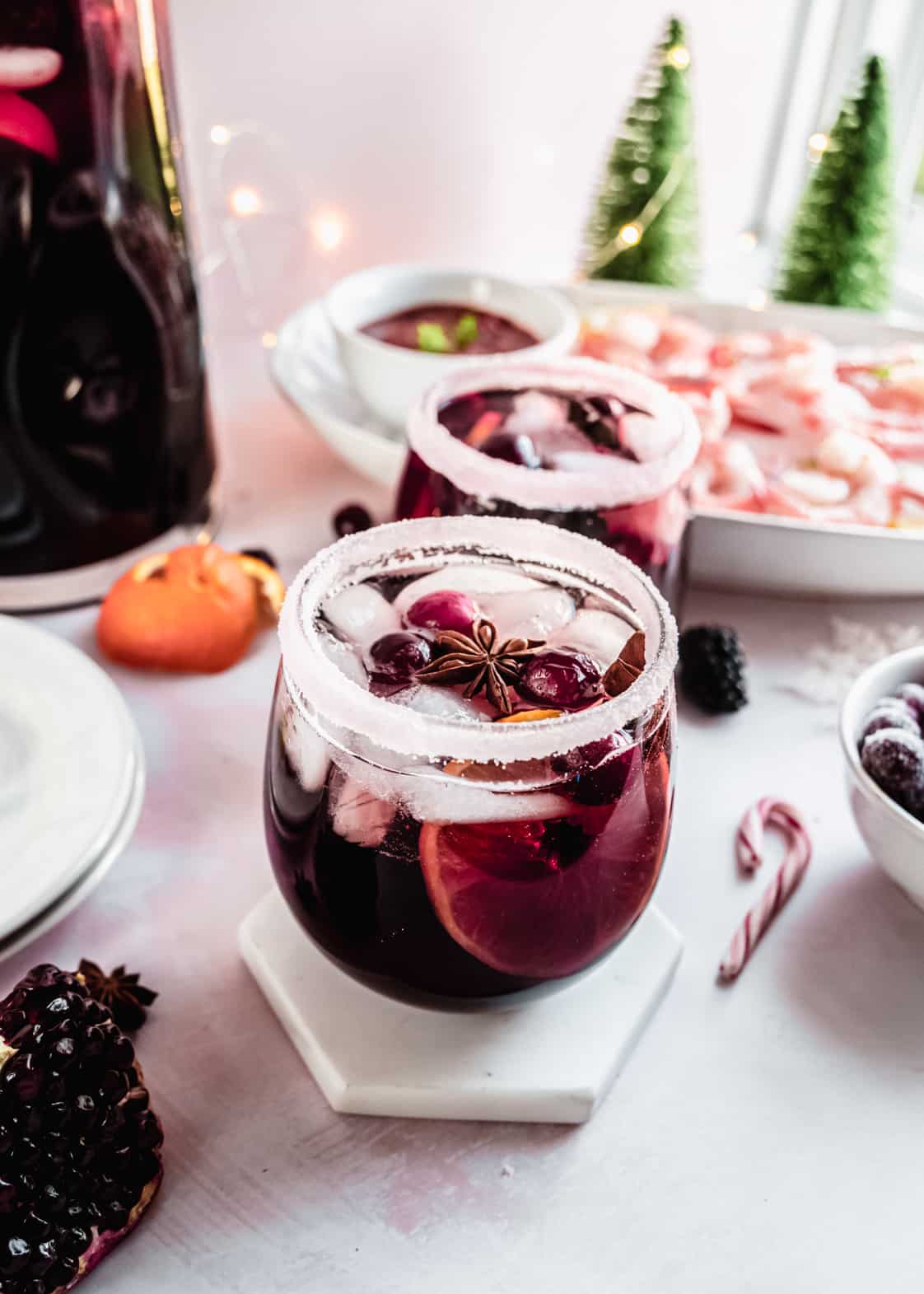 A festive glass of Christmas sangria filled with ice, citrus slices, cranberries, and star anise, served in a sugar-rimmed glass, with holiday décor, shrimp cocktail, and winter greenery in the background.