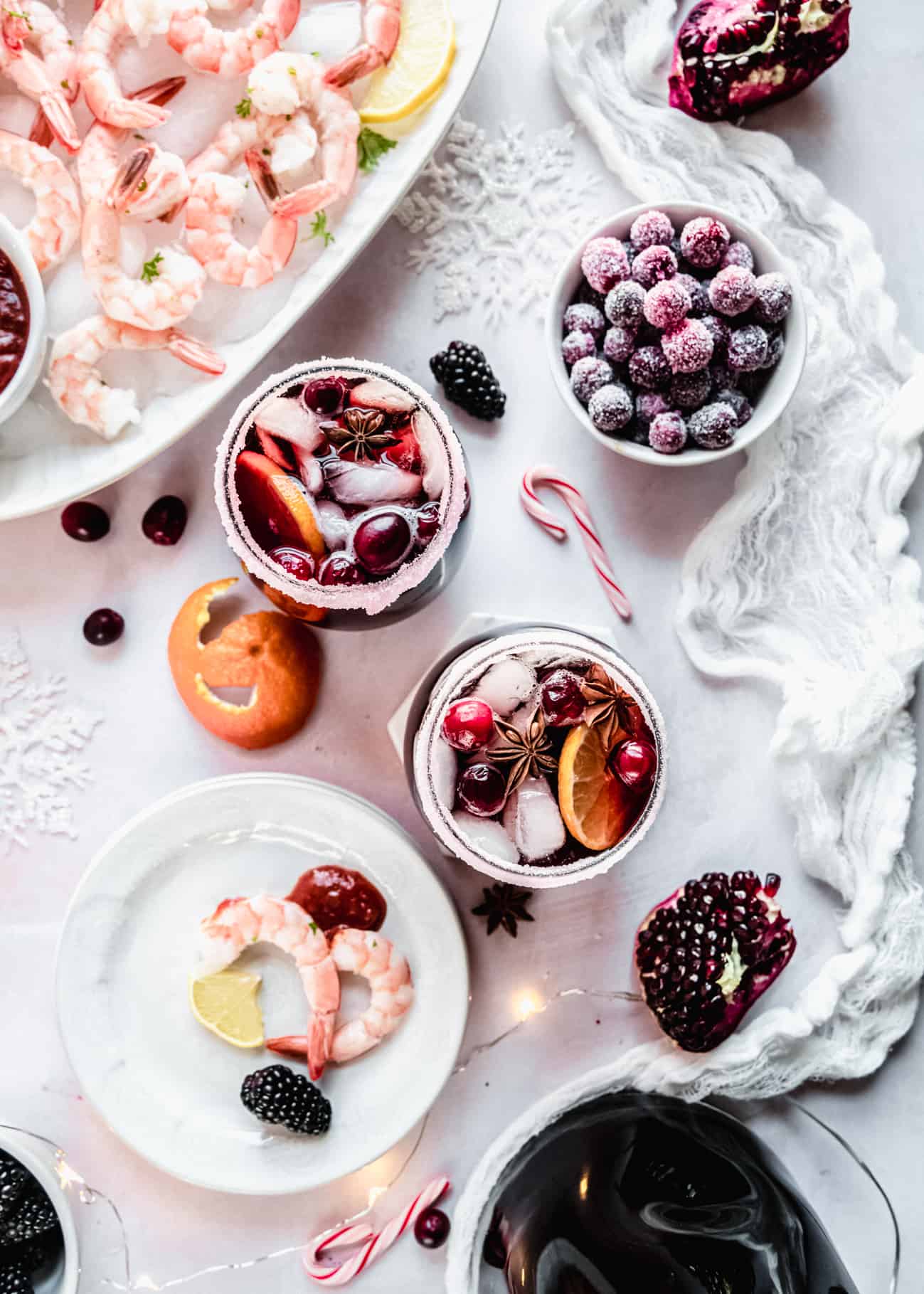 A festive holiday spread featuring two glasses of Christmas sangria garnished with citrus slices, cranberries, ice, and star anise, surrounded by sugared cranberries, shrimp cocktail, candy canes, and winter fruit on a bright, snowy-inspired tabletop.