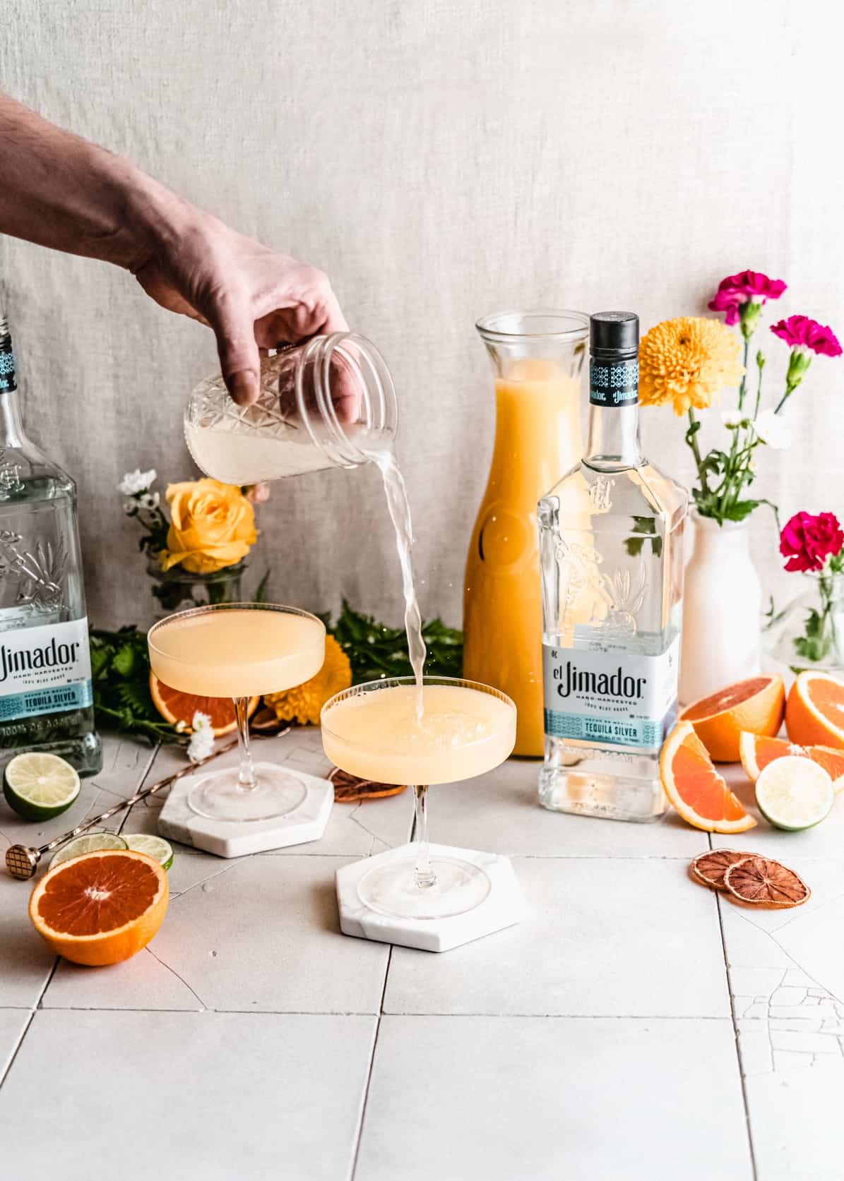 Ginger beer being poured from cocktail shaker into cocktail glass.  Tequila bottle, orange juice and flowers in the background