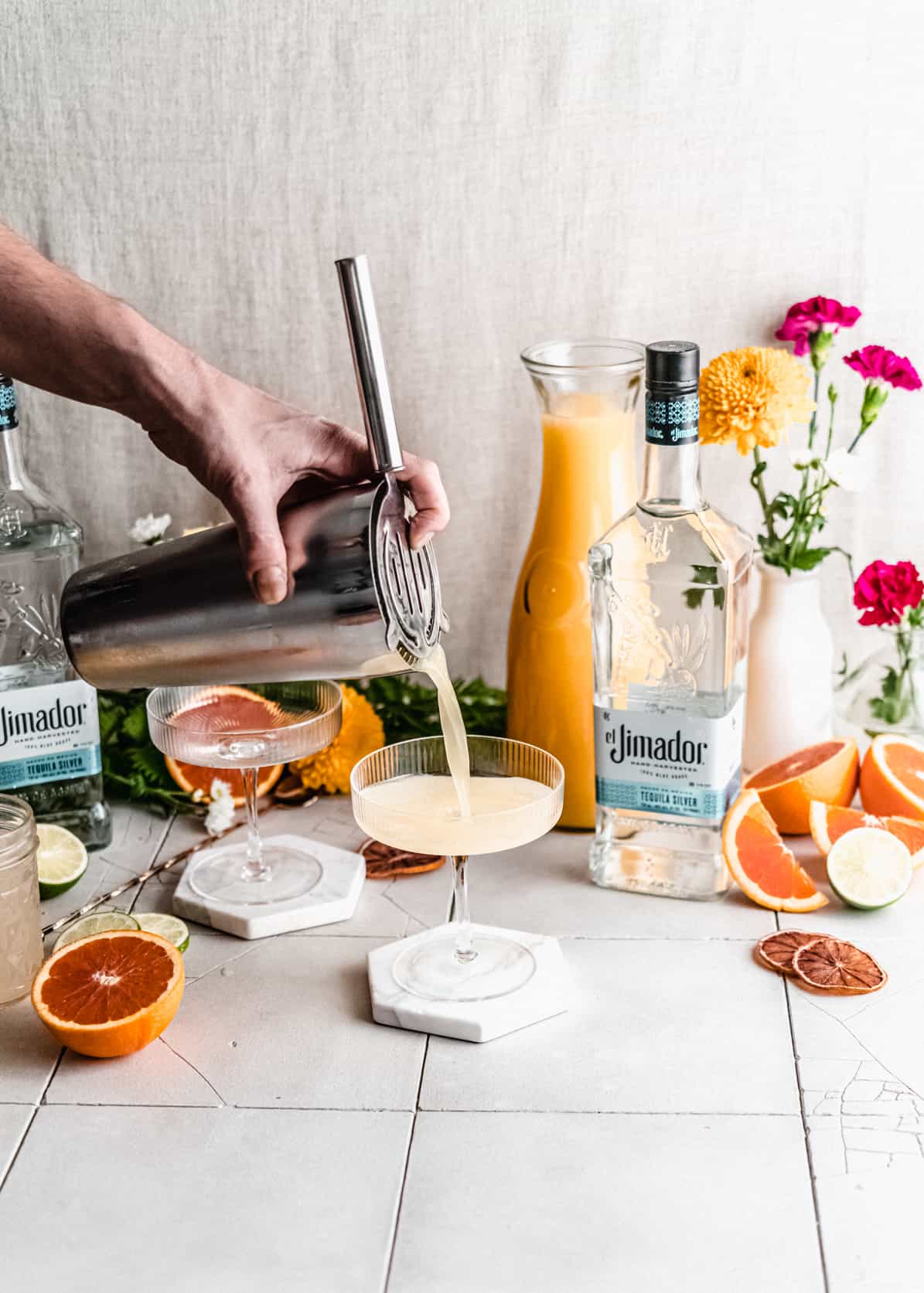 tequila spritzer being poured from cocktail shaker into cocktail glass.  Tequila bottle, orange juice and flowers in the background