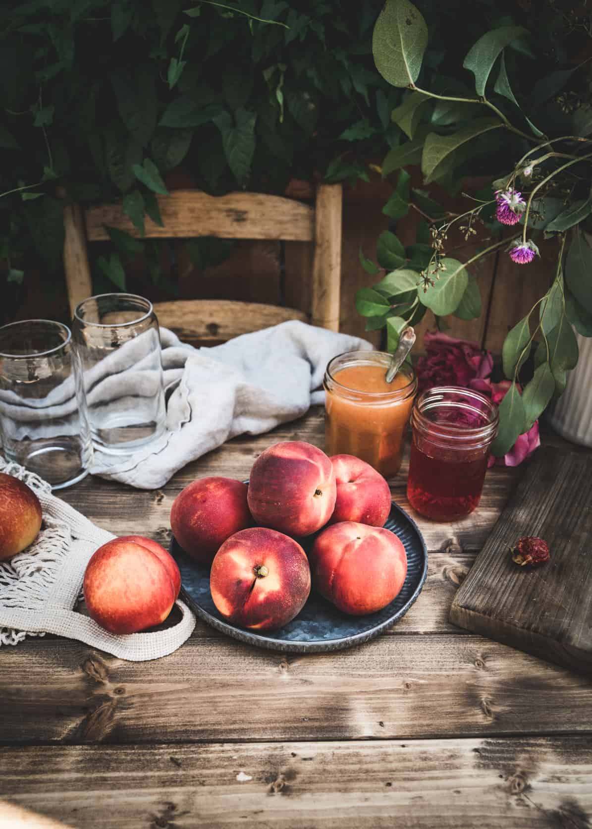 Whole fresh peaches on metal plate on wood table.  Peach puree and peach simple syrup are in glass jars behind the plate, and empty glasses are on the table.  Greenery surrounds the table. 