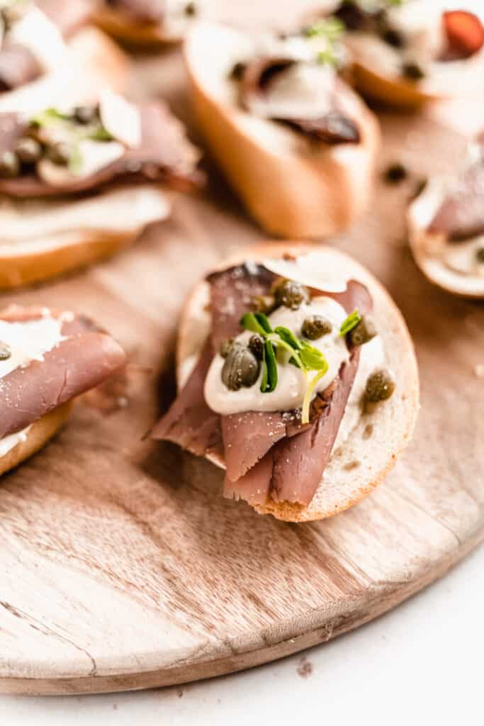 close up shot of roast beef crostini garnished with horseradish dijon sauce, capers and microgreens on a wood serving platter