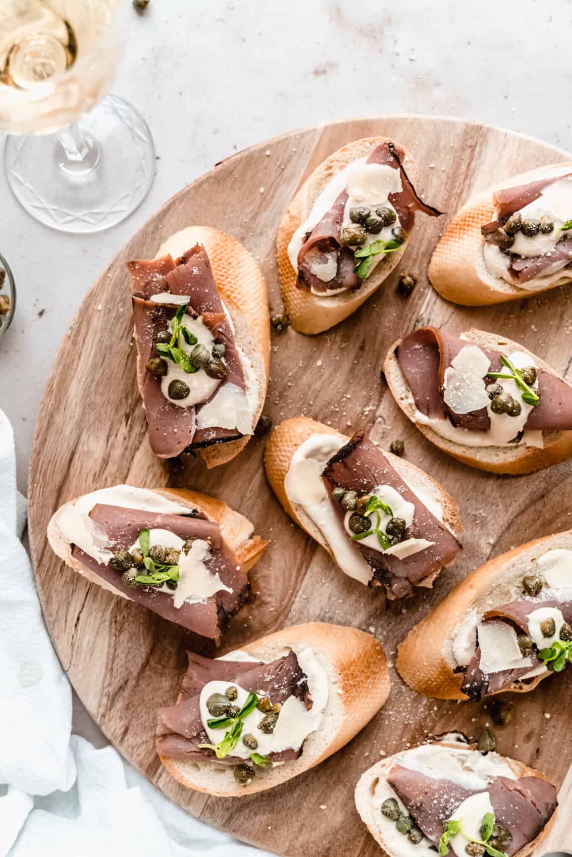 overhead shot of roast beef crostini garnished with horseradish dijon sauce, capers and microgreens on a wood serving platter, placed next to a glass of wine 