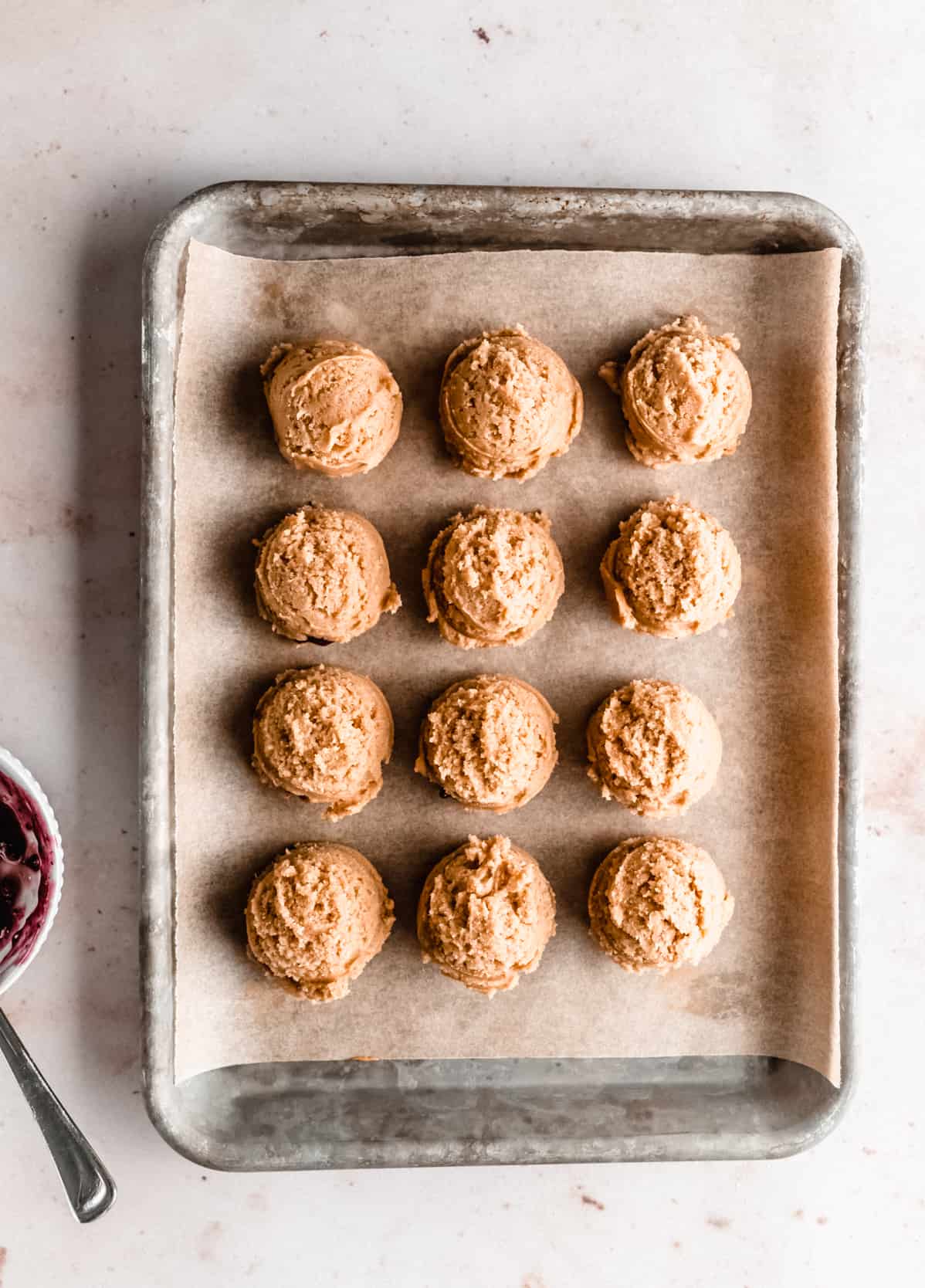 Scoops of peanut butter cookie dough lined up on a parchment-lined baking sheet, ready to be filled with jelly to make peanut butter and jelly cookies. A small bowl of dark red jelly and a spoon sit nearby.