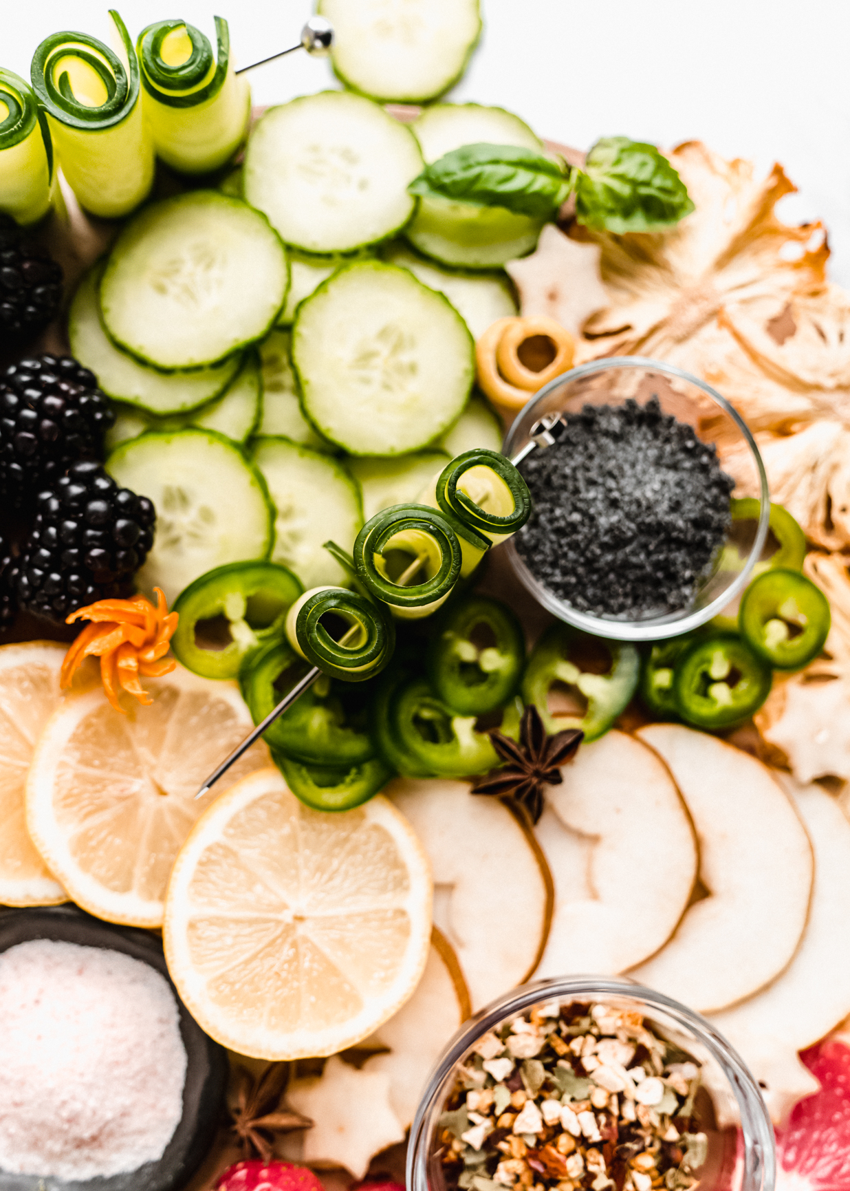 close up of sliced cucumber, blackberries, fresh lemon on cocktail garnish board 