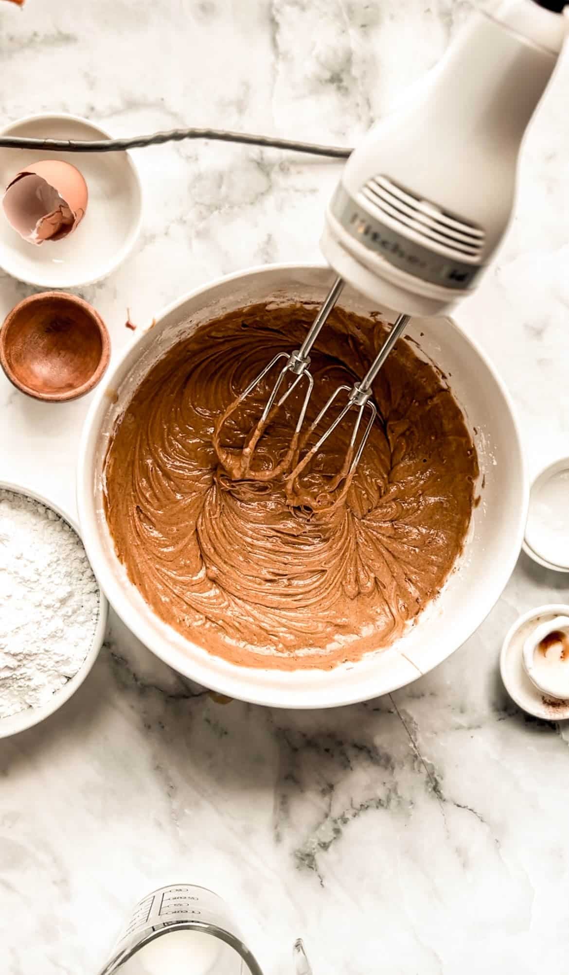 A mixing bowl filled with smooth gingerbread snack cake batter sits on a marble surface, with an electric hand mixer resting in the bowl. Surrounding ingredients like flour, an egg shell, and spices show the cake-making process.