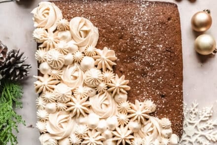 A gingerbread snack cake decorated on one side with intricate piped buttercream swirls and stars, surrounded by holiday greenery, ornaments, snowflakes, and a small gingerbread cookie.