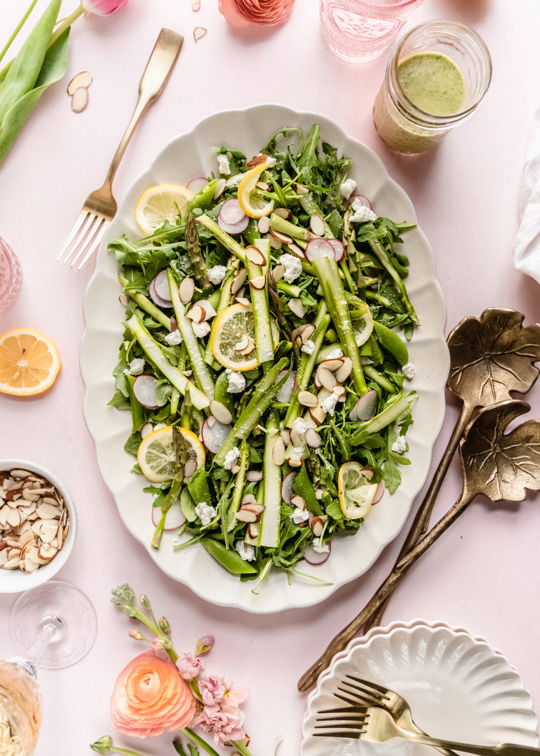 Bright overhead tablescape featuring a raw asparagus salad with shaved asparagus ribbons, arugula, sugar snap peas, radishes, goat cheese, sliced almonds, and lemon slices, served on a white platter with lemon basil dressing, gold utensils, fresh flowers, and glasses on a soft pink background.