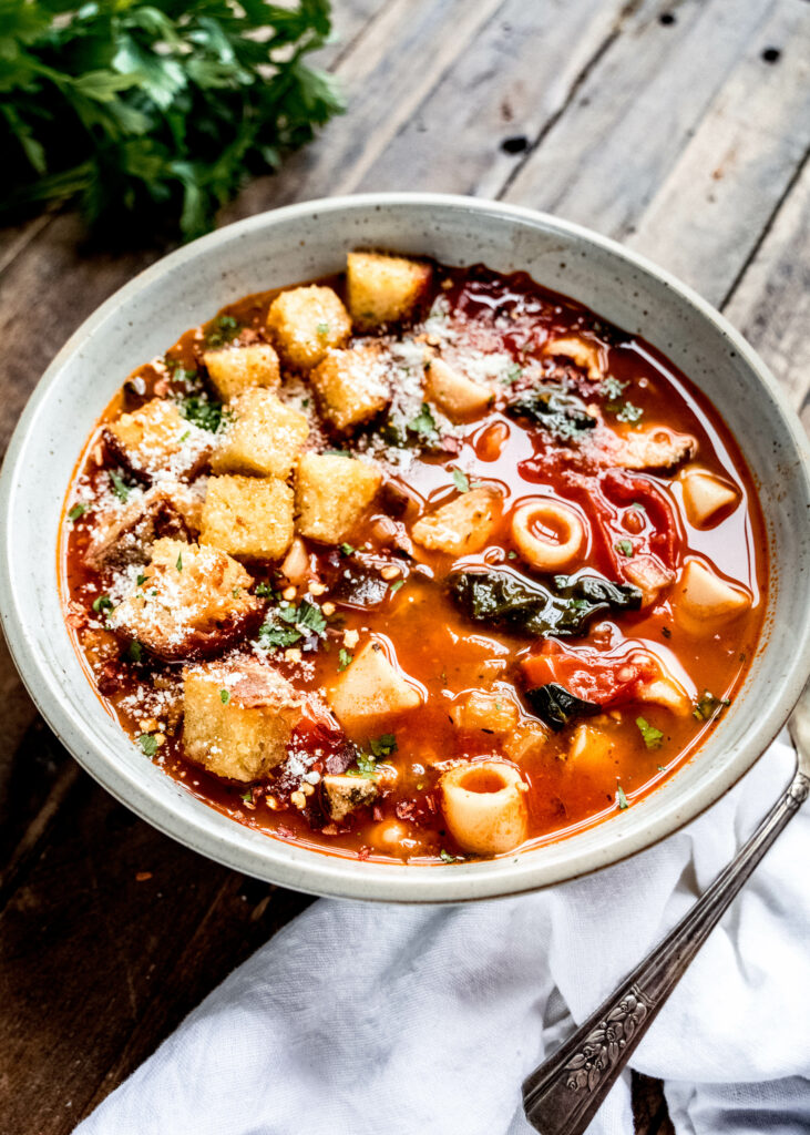 45 degree angle of instant pot minestrone soup garnished with fresh herbs, grated parmesan and croutons with a white napkin and spoon next to the bowl laying on a wood surface 