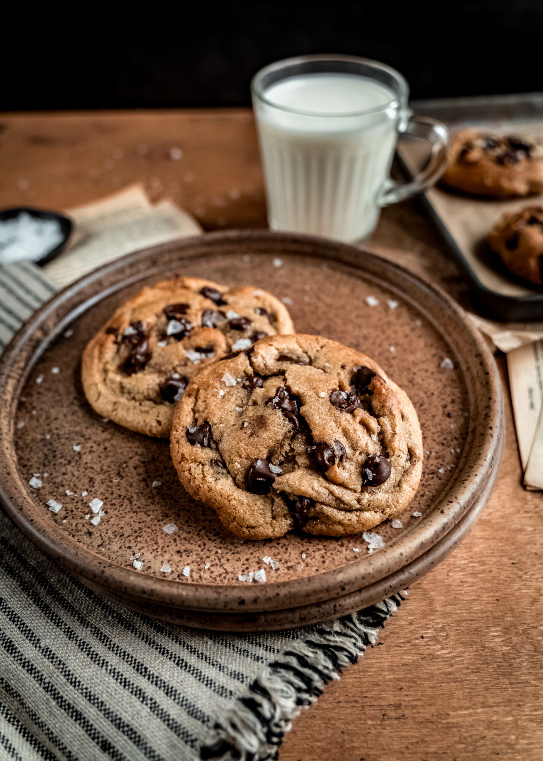 Side shot of two chocolate chip cookies on brown plate with a striped napkin, a glass of milk and a cookie sheet with baked chocolate chip cookies in the background 