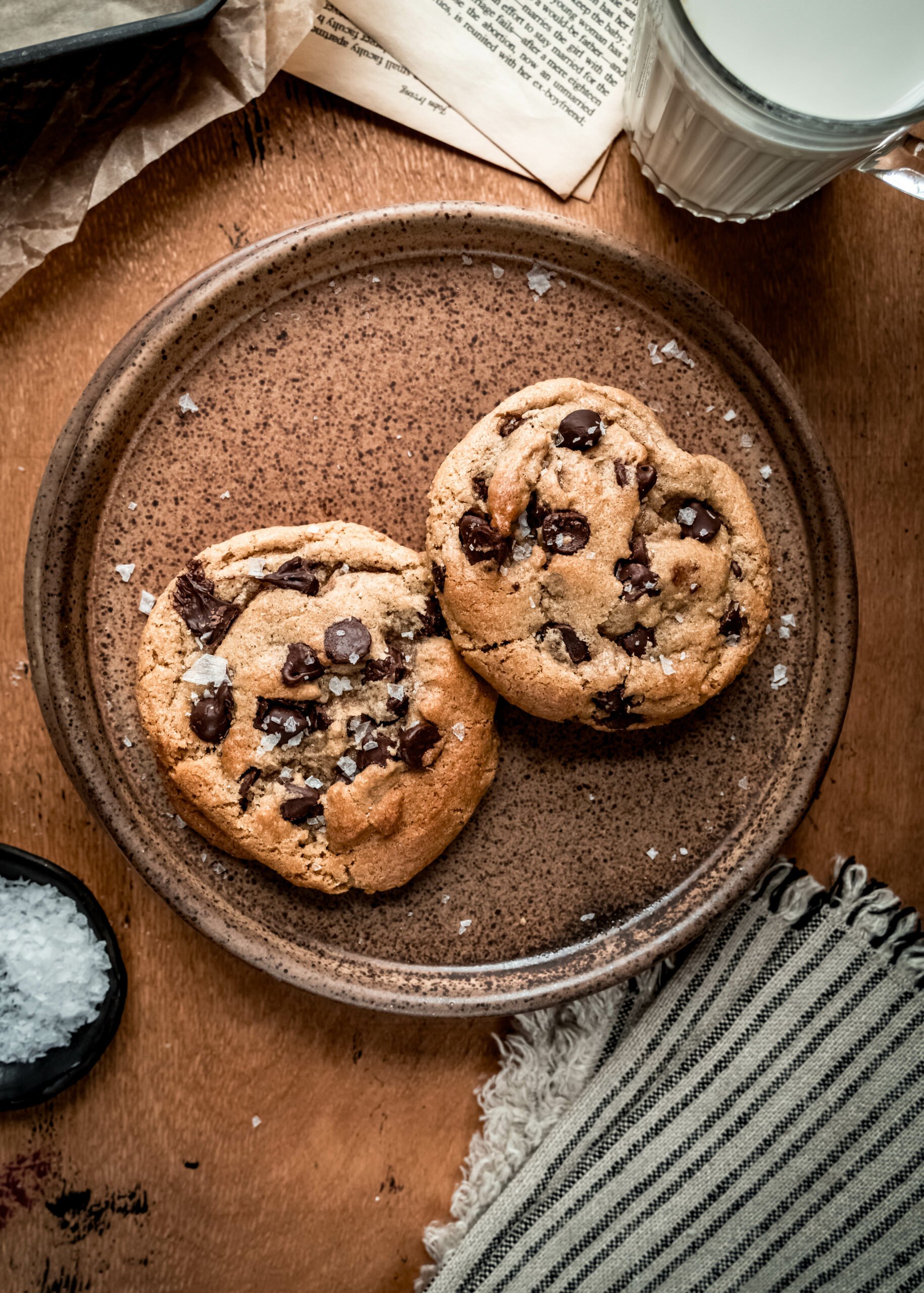 overhead shot of two chocolate chip cookies on brown plate with a striped napkin, a glass of milk 