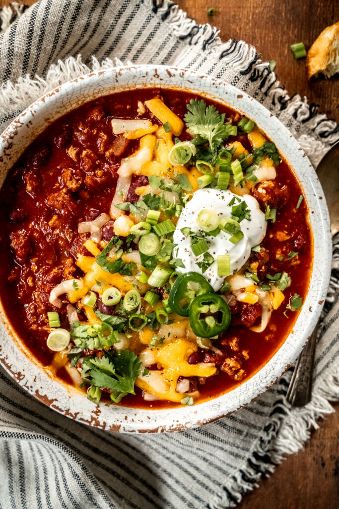bowl of turkey chili garnished with shredded cheese, cilantro, sliced green onion and sour cream.  The bowl is sitting on a striped napkin on a wood table. 